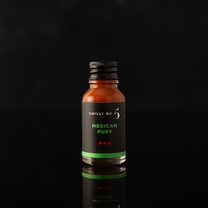 May include: A small glass bottle of hot sauce with a black label that says "CHILLI No. 5 MEXICAN FURY" with three red dots underneath the text. The bottle is filled with a red-brown liquid and has a black screw-top lid.