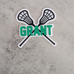 May include: A white sticker with two black lacrosse sticks crossed over each other. The word "GRANT" is written in green letters on the sticker.
