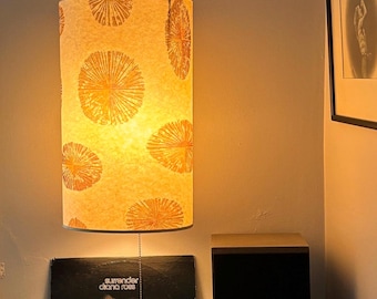 May include: A hanging cylindrical lampshade with a repeating orange floral pattern illuminates a room. Below, a vintage album cover, a small potted plant, and a dark sculpture sit on a wooden shelf. A framed artwork hangs on the wall.