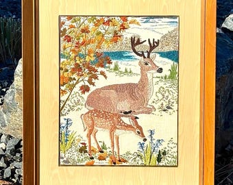 May include: A framed embroidery art piece featuring a buck and a fawn in a landscape with autumn foliage, a blue lake, and green grass. The artwork is enclosed in a light-coloured mat and a medium-toned wooden frame with a smooth, polished finish.