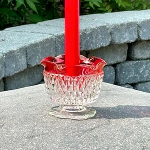 May include: A red candle in a clear glass candle holder with a red, ruffled rim. The candle holder has a diamond-cut pattern. The background features green foliage and a grey stone wall.