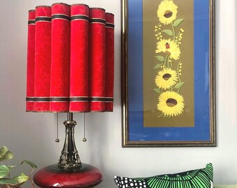 May include: A table lamp with a red pleated fabric shade featuring horizontal black and white stripes, set on a dark red and bronze-toned base. Beside it hangs a framed vertical artwork depicting four yellow sunflowers on a brown background.