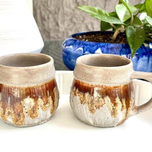 May include: Two handmade ceramic mugs with a rustic design. The mugs have a light beige top and handle, with a brown and gold glaze that drips down the sides. The mugs are sitting on a white surface.