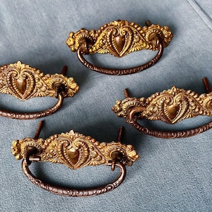 May include: Four antique brass drawer pulls with ornate, heart-shaped designs. The pulls have a gold finish with dark brown handles and mounting posts. The hardware is arranged on a light blue surface.