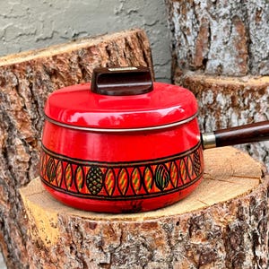 Peut inclure: Une casserole en métal émaillé rouge vif avec un couvercle assorti et un long manche en bois brun foncé. La marmite présente une bande décorative autour du centre avec des formes ovales répétitives en noir, jaune et vert. Le couvercle a un bouton brun.