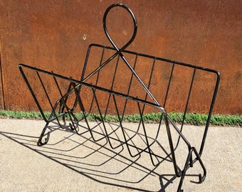 May include: Black metal magazine rack with a unique curved design. The rack has a series of vertical bars for holding magazines or newspapers.