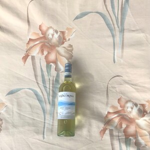 May include: A bottle of Mezzacorona Pinot Grigio wine on a white background with a floral pattern. The bottle is clear glass with a white label and a gold screw top.