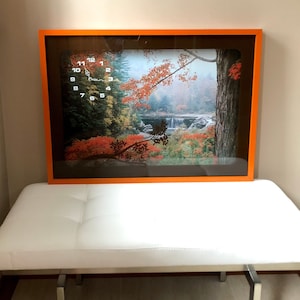 Peut inclure: Une impression encadrée d'un paysage d'automne avec une cascade et des arbres aux feuilles rouges. Le cadre est orange et l'impression est derrière une vitre.