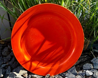 May include: A bright orange ceramic plate with a slightly raised rim. The plate is sitting on a bed of gray gravel.