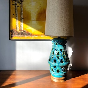 Peut inclure: Une lampe en céramique bleu turquoise avec un motif géométrique et un abat-jour en tissu blanc. La lampe est posée sur une table en bois devant une peinture encadrée d'un paysage.