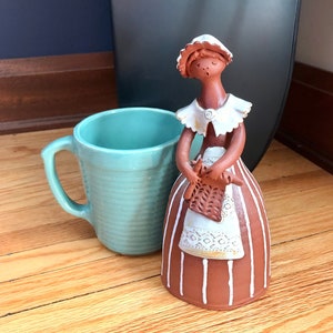 May include: A ceramic figurine of a woman wearing a white dress with brown stripes and a white collar. She is holding a brown and white knitted object in her hands. The figurine is standing on a wooden surface.