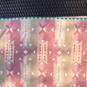 Könnte beinhalten: Ein Stoff mit einem Muster im Südwest-Stil in Rosa, Beige und Hellblau. Das sich wiederholende Muster umfasst geometrische Formen und stilisierte Motive, mit einem türkisfarbenen Rand. Der Stoff scheint aus Baumwolle oder einem ähnlichen Material zu sein.