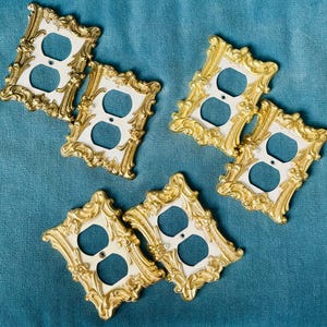 May include: Ornate, gold-colored electrical outlet covers with a white center. The covers have a decorative, vintage-inspired design. The outlets are arranged on a teal-colored surface.