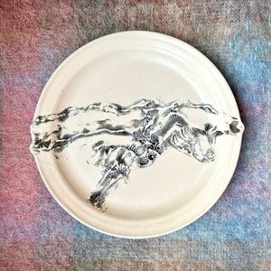 May include: A white ceramic plate featuring a black and white abstract design. The design resembles a flowing wave or a mountain range.