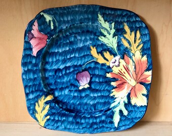 May include: A blue ceramic square plate with a floral design. The plate features a large poppy flower with yellow, orange, and red petals, and green leaves. The background is a textured blue.