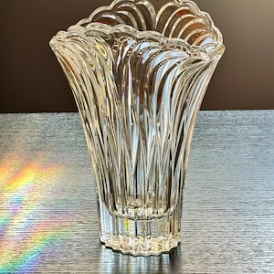 May include: A clear glass vase with a fluted design. The vase has a wide, scalloped top and a flared shape. Vertical ridges create a textured effect. The vase is on a dark surface, with a rainbow prism effect visible.