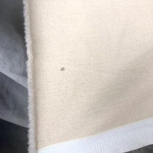 May include: A close-up of a cream-colored fabric with a white trim. The fabric has a subtle texture and a small brown spot.