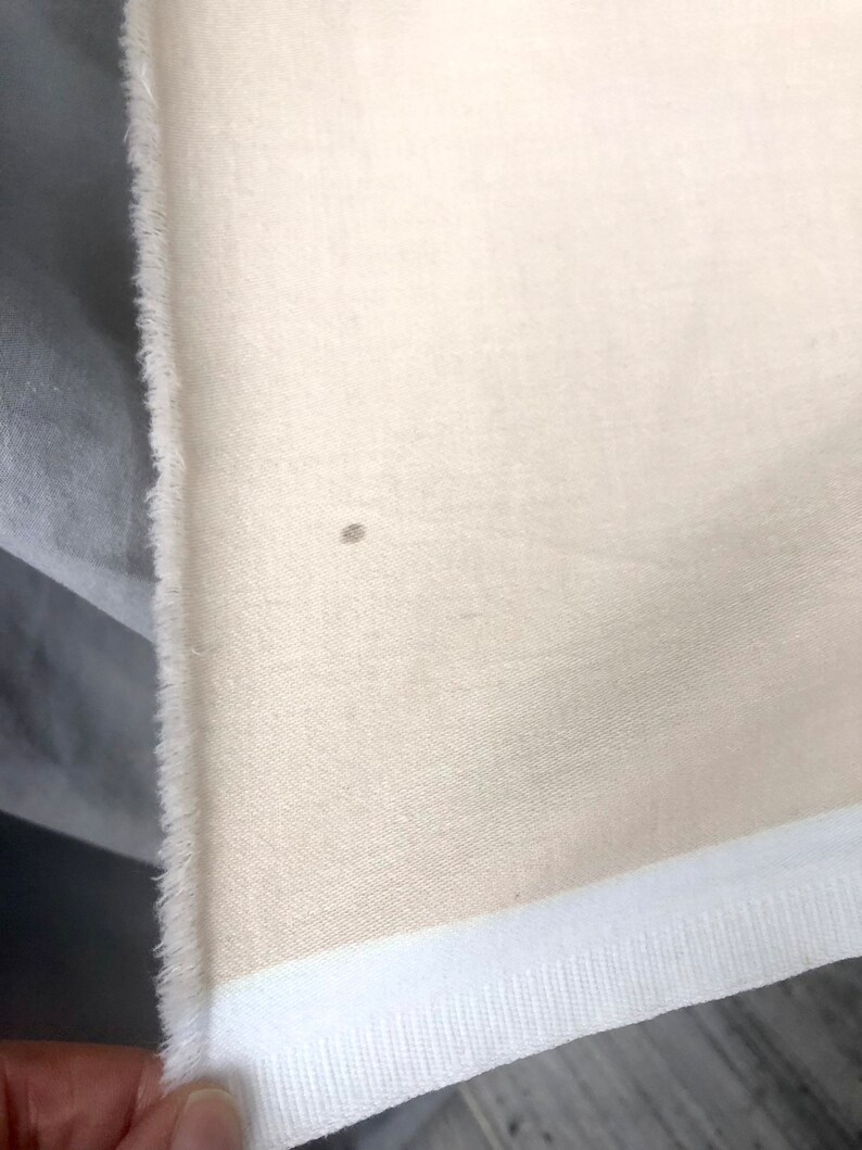 May include: A close-up of a cream-colored fabric with a white trim. The fabric has a subtle texture and a small brown spot.
