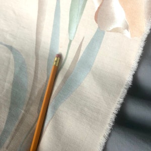 May include: A close-up of a cream-colored fabric with a floral pattern. The fabric has a subtle texture and a frayed edge. A pencil is lying on top of the fabric.