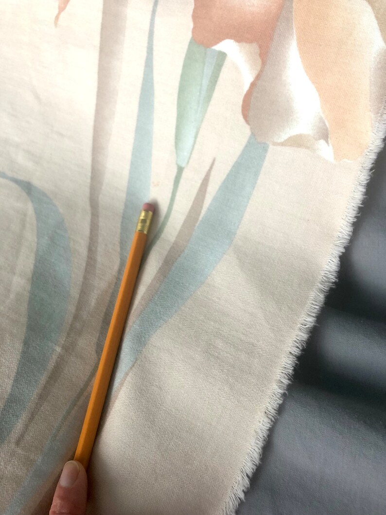 May include: A close-up of a cream-colored fabric with a floral pattern. The fabric has a subtle texture and a frayed edge. A pencil is lying on top of the fabric.