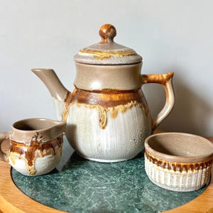 May include: A ceramic tea set featuring a teapot with a lid, a creamer, and a small bowl. The set has a beige, brown, and white color scheme with a drip glaze design. The teapot has a handle and spout.