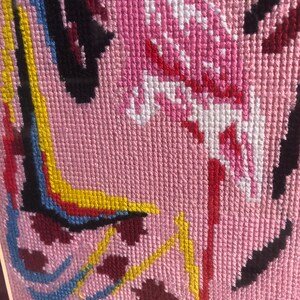 May include: Close-up of a pink cross-stitch embroidery featuring colourful geometric shapes and a pink and white bird design.
