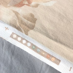May include: A close-up of a beige fabric with a subtle floral pattern. The fabric has a textured surface and a white border with a row of small, colorful circles.