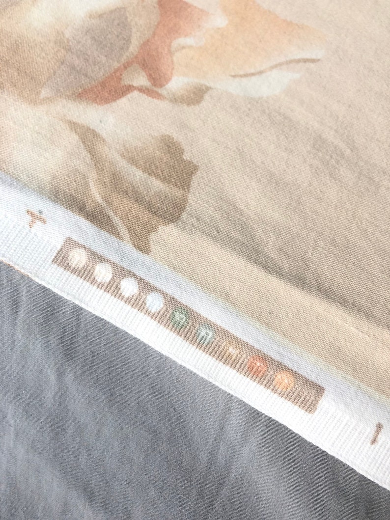 May include: A close-up of a beige fabric with a subtle floral pattern. The fabric has a textured surface and a white border with a row of small, colorful circles.