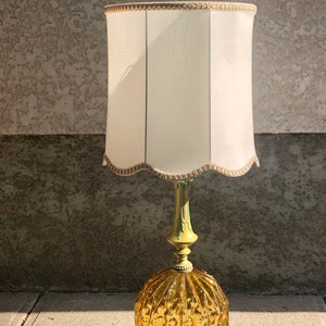 Pu&ograve; includere: Una lampada da tavolo con una base rotonda in vetro color ambra caratterizzata da un motivo a coste. La base poggia su un supporto in metallo verde chiaro. &Egrave; completata da un alto paralume cilindrico bianco sporco con bordo smerlato e finiture oro.