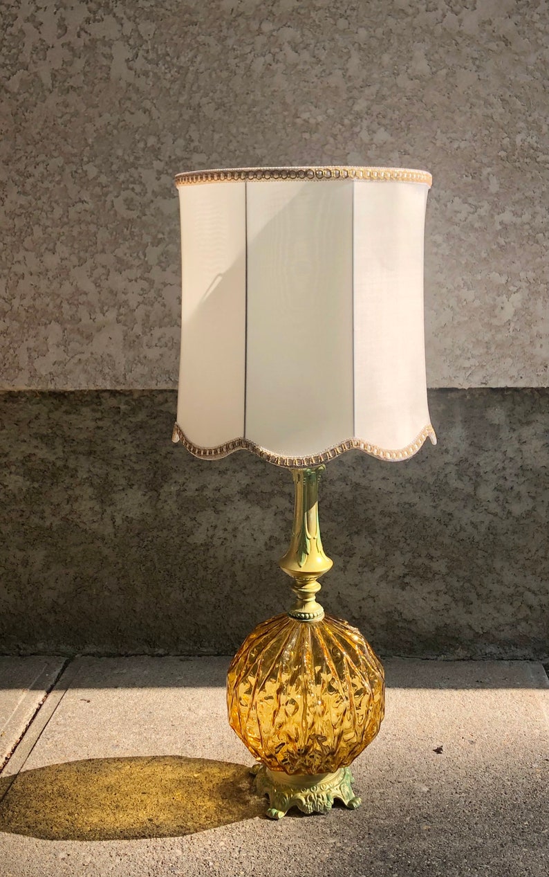Pu&ograve; includere: Una lampada da tavolo con una base rotonda in vetro color ambra caratterizzata da un motivo a coste. La base poggia su un supporto in metallo verde chiaro. &Egrave; completata da un alto paralume cilindrico bianco sporco con bordo smerlato e finiture oro.