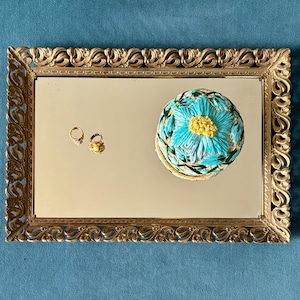 May include: A gold-framed mirror with a decorative floral design. The mirror reflects a small, round, blue and yellow floral-patterned box and two small rings. The rings are gold with gemstones and floral details.