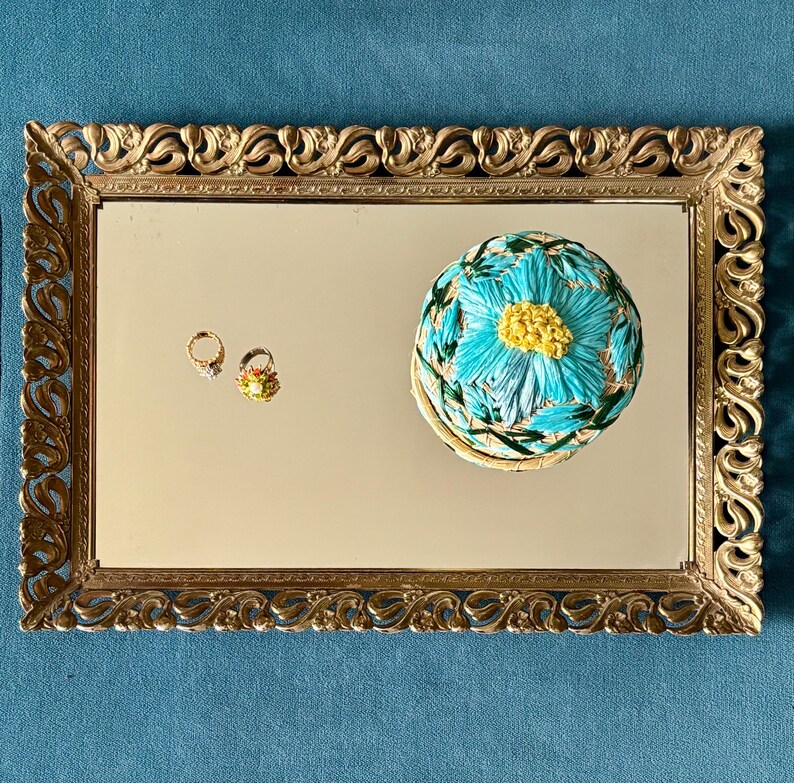May include: A gold-framed mirror with a decorative floral design. The mirror reflects a small, round, blue and yellow floral-patterned box and two small rings. The rings are gold with gemstones and floral details.