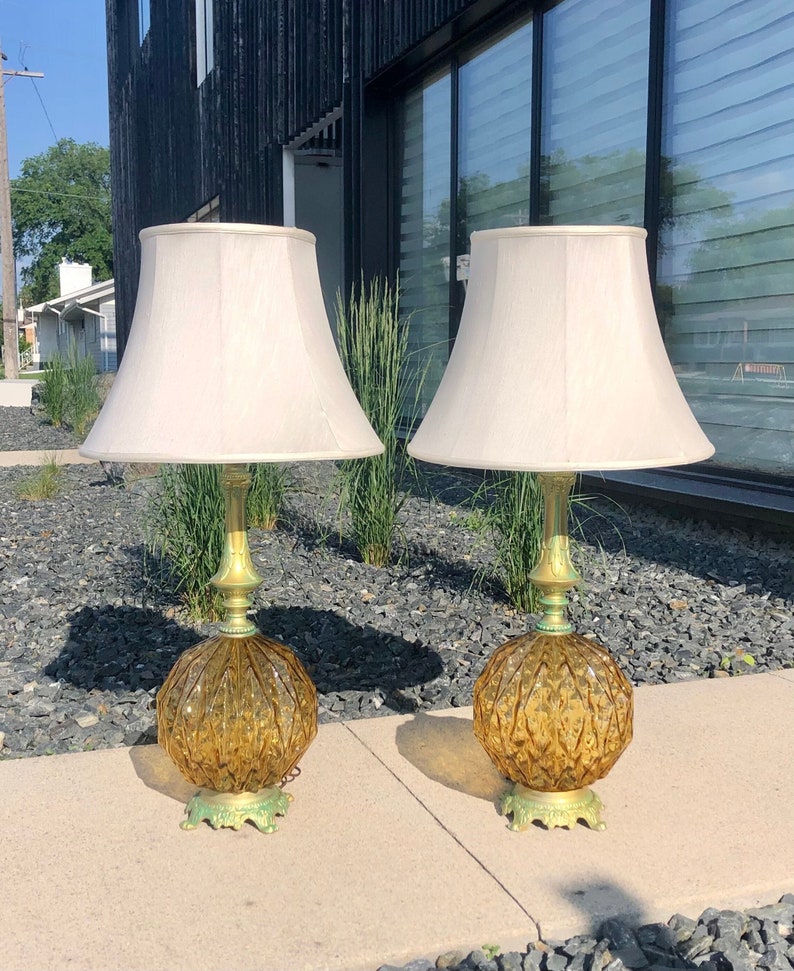 Pu&ograve; includere: Una coppia di lampade da tavolo con basi in vetro color ambra a motivo diamantato e accenti verde metallizzato. Ogni lampada presenta uno stelo decorativo sottile e un ampio paralume in tessuto bianco sporco dalla forma a campana.