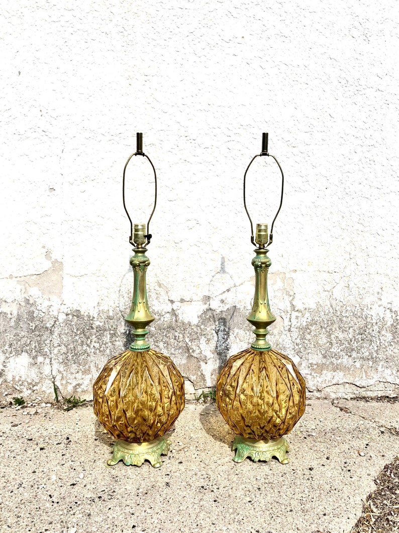 Pu&ograve; includere: Una coppia di lampade da tavolo con basi in vetro color ambra a motivo diamantato. Ogni lampada presenta un collo e una base in metallo dai toni verdi con dettagli decorati, sormontati da un'arpa in metallo e un portalampada standard.