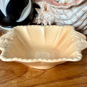 Puede incluir: Un plato de cerámica vintage de color melocotón claro, con un borde festoneado y adornos ornamentados. El plato tiene un diseño de sol en el centro y está colocado sobre una superficie de madera. Hay objetos decorativos en el fondo.