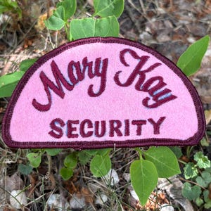 May include: A pink, semi-circular patch with the words "Mary Kay" in cursive and "SECURITY" in block letters. The text is a dark burgundy colour, outlined in a darker burgundy, and set against a pink background. The patch is surrounded by a burgundy border.
