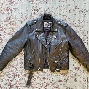 May include: A black leather motorcycle jacket with a zipper closure and multiple pockets. The jacket has a label that reads "First" on the left side of the chest.