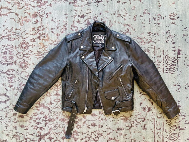 May include: A black leather motorcycle jacket with a zipper closure and multiple pockets. The jacket has a label that reads "First" on the left side of the chest.