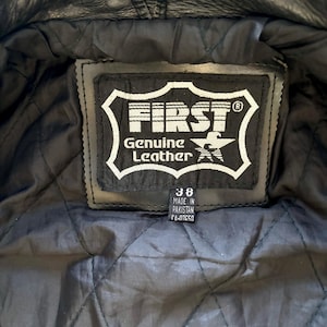 May include: Close-up of a black leather jacket's inner lining with a black label that reads "FIRST Genuine Leather" and a star logo. The label also includes the size "38" and "Made in Pakistan" text.
