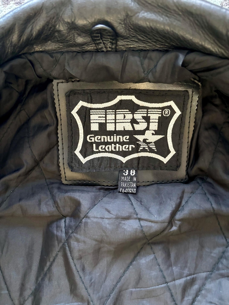 May include: Close-up of a black leather jacket's inner lining with a black label that reads "FIRST Genuine Leather" and a star logo. The label also includes the size "38" and "Made in Pakistan" text.
