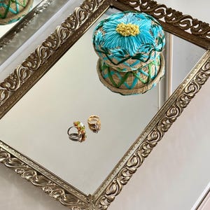 May include: A gold-coloured rectangular tray with detailed, ornate edges, reflecting a blue and yellow floral pincushion and two rings. The pincushion is round, with a woven base and a bright blue flower design. The rings have colourful gemstones.