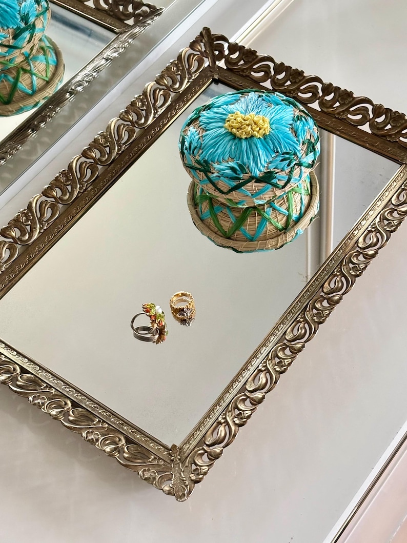 May include: A gold-coloured rectangular tray with detailed, ornate edges, reflecting a blue and yellow floral pincushion and two rings. The pincushion is round, with a woven base and a bright blue flower design. The rings have colourful gemstones.