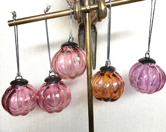 Könnte beinhalten: Ein dekorativer Messingständer mit einem vogelartigen Design, der fünf Glasornamente hält. Die Ornamente sind in verschiedenen Rosa- und Orangetönen gehalten und haben silberfarbene Kappen und Aufhängeschnüre. Die Ornamente haben eine gerippte Textur.