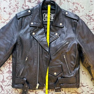 May include: Black leather motorcycle jacket with a zipper closure, two front pockets, and a belt at the waist. The jacket has a label that reads "First Genuine Leather".