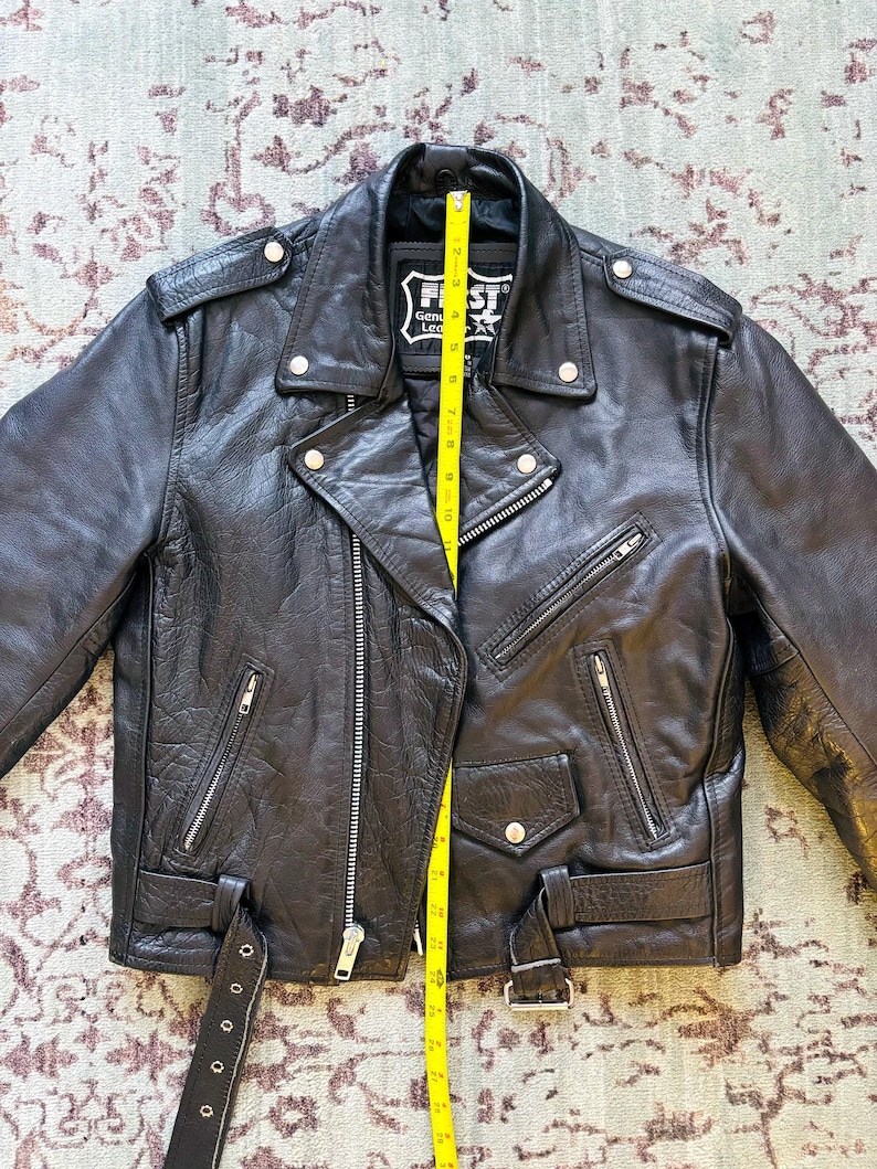 May include: Black leather motorcycle jacket with a zipper closure, two front pockets, and a belt at the waist. The jacket has a label that reads "First Genuine Leather".