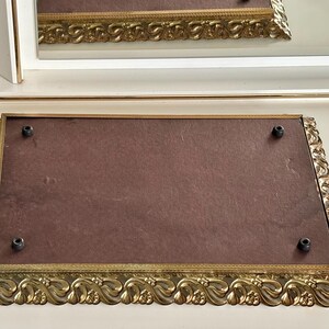 May include: An ornate, rectangular gold-toned tray with a dark brown interior. The tray features a decorative, scrolling edge and four black screw heads. A white and gold-trimmed mirror is in the background.