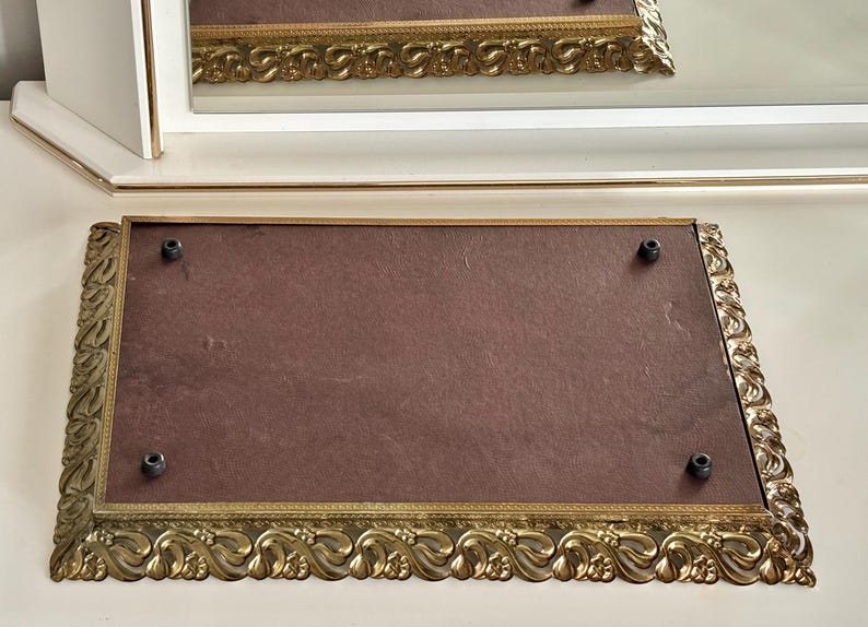 May include: An ornate, rectangular gold-toned tray with a dark brown interior. The tray features a decorative, scrolling edge and four black screw heads. A white and gold-trimmed mirror is in the background.