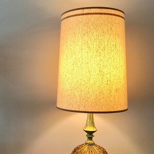 Pu&ograve; includere: Una lampada da tavolo con base in vetro ambrato testurizzato, supporto in metallo dorato e paralume cilindrico beige con bordo marrone. Accanto alla lampada si trova un telefono a disco arancione brillante con cavo e tastiera neri.