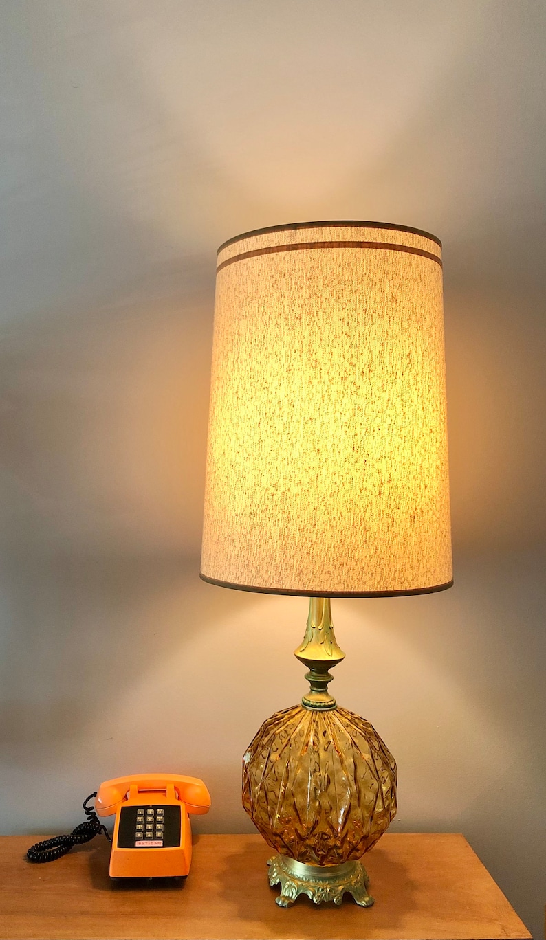 Pu&ograve; includere: Una lampada da tavolo con base in vetro ambrato testurizzato, supporto in metallo dorato e paralume cilindrico beige con bordo marrone. Accanto alla lampada si trova un telefono a disco arancione brillante con cavo e tastiera neri.