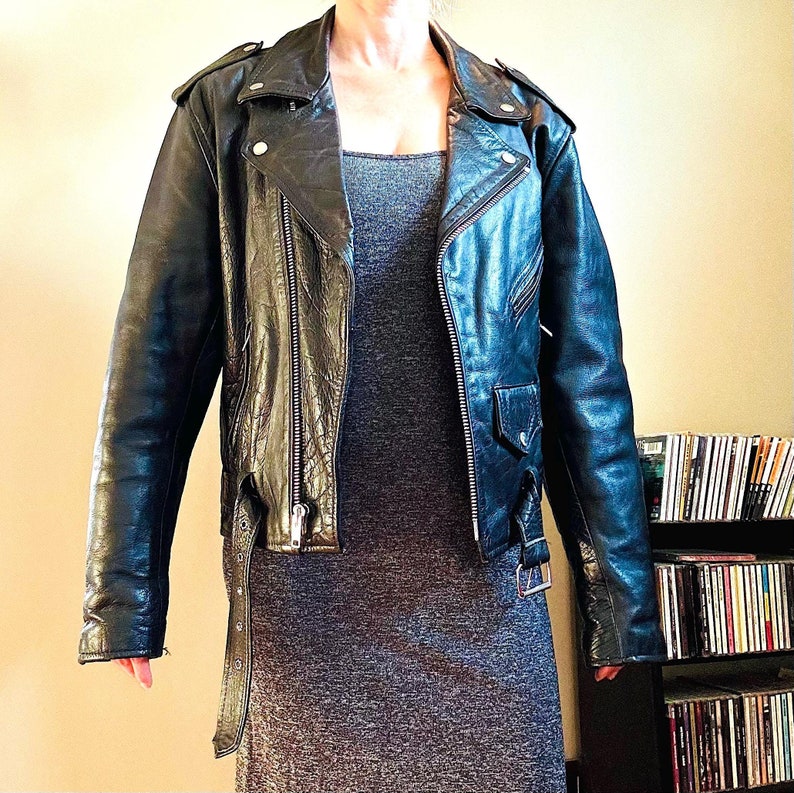 May include: A black leather motorcycle jacket with a zipper closure and multiple pockets. The jacket has a classic biker style and features a silver zipper and snaps.